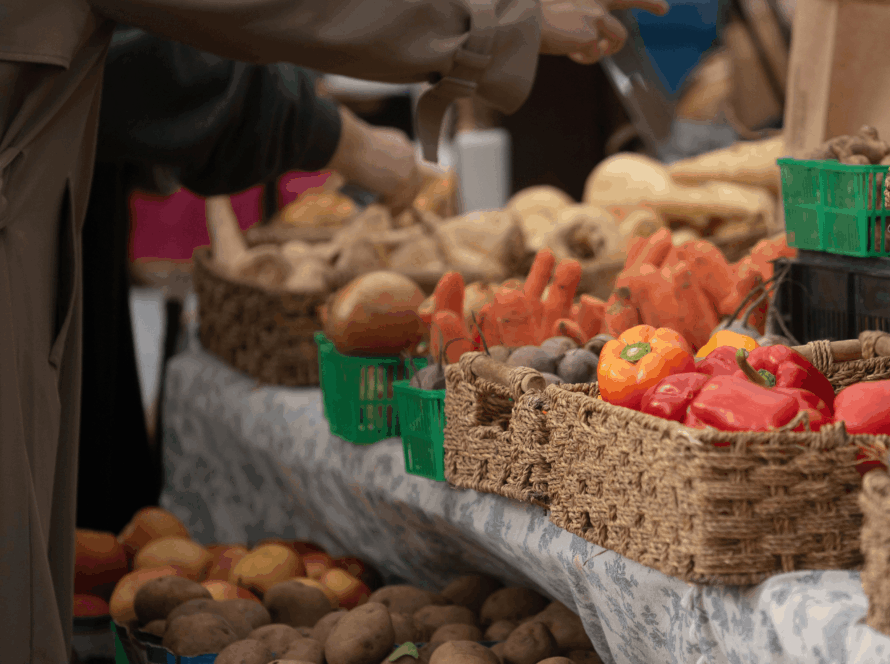 Person in a tan coat reaches to pick potatoes from crates at a market stall, with baskets of root vegetables in the foreground